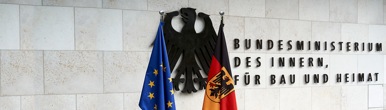 Im Außenbereich des BMI stehen vor dem Bundesadler und dem Schriftzug des Ministeriums ene EU- und Bundesflagge Im Außenbereich des BMI stehen vor dem Bundesadler und dem Schriftzug des Ministeriums ene EU- und Bundesflagge