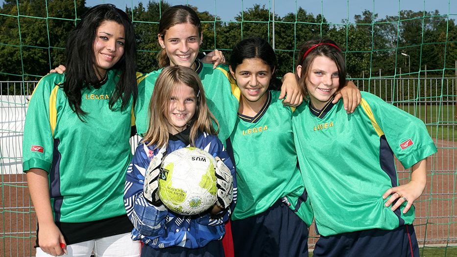 Mädchen verschiedener Nationalitäten (bekleidet in Fussballtrikots) stehen in einem Fussballtor und lächeln fröhlich in die Kamera