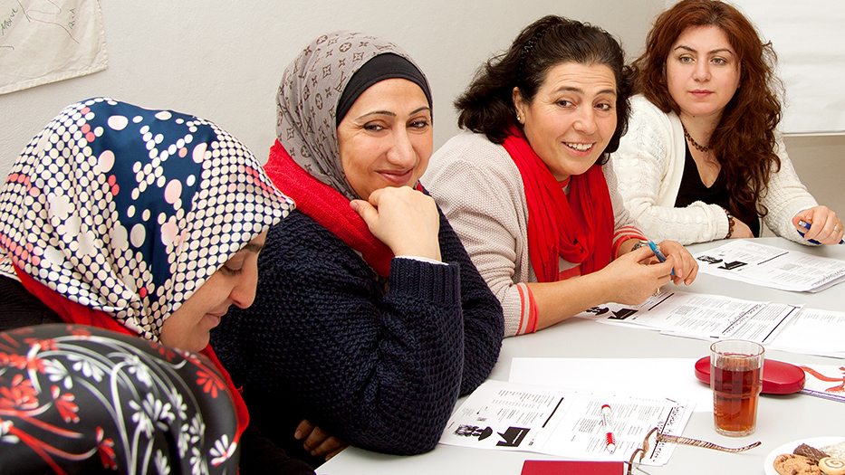 Vier Frauen, zwei davon tragen ein Kopftuch, sitzen an einem Tisch, vor ihnen ausgebreitete Arbeitsblätter