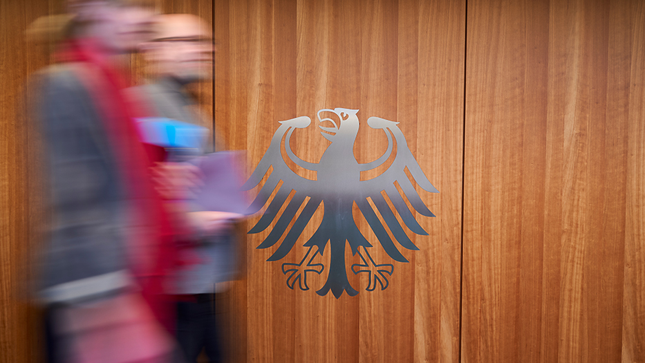 Unscharf fotografierte Personen laufen an grauem Bundesadler auf einer Holzwand vorbei