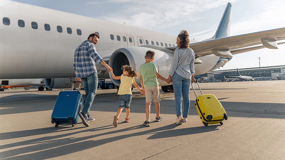 Das Foto zeigt lächelnde Eltern und zwei Kinder. Die Familie geht mit Koffern auf einem Rollfeld in Richtung eines großen Flugzeugs.
