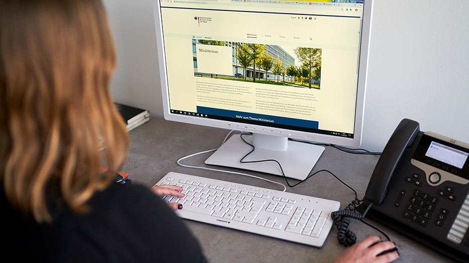 Von hinten fotografierte Frau arbeitet am Computer, auf dem Monitor die Webseite des BMI