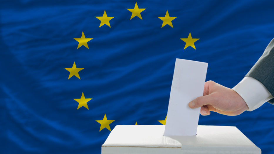 Ein Wahlzettel wird in eine Wahlurne gesteckt mit einer Europaflagge im Hintergrund