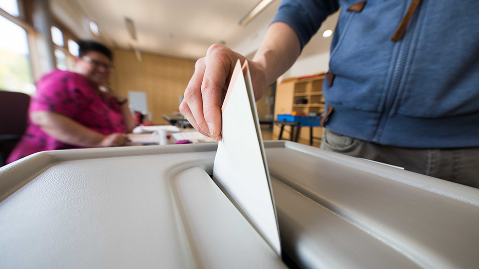 Eine Hand wirft einen Wahlzettel in eine Urne, im Hintergrund unscharf eine am Tisch sitzende Frau 