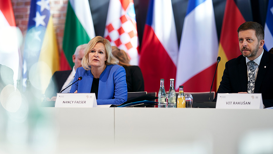 Bundesinnenministerin Nancy Faeser sits in front of a microphone, behind her flags of the countries participating in the Western Balkans Conference
