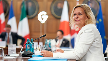 Federal Minister of the Interior Nancy Faeser sits at a conference table; other people, flags and the G7 logo are blurred in the background.